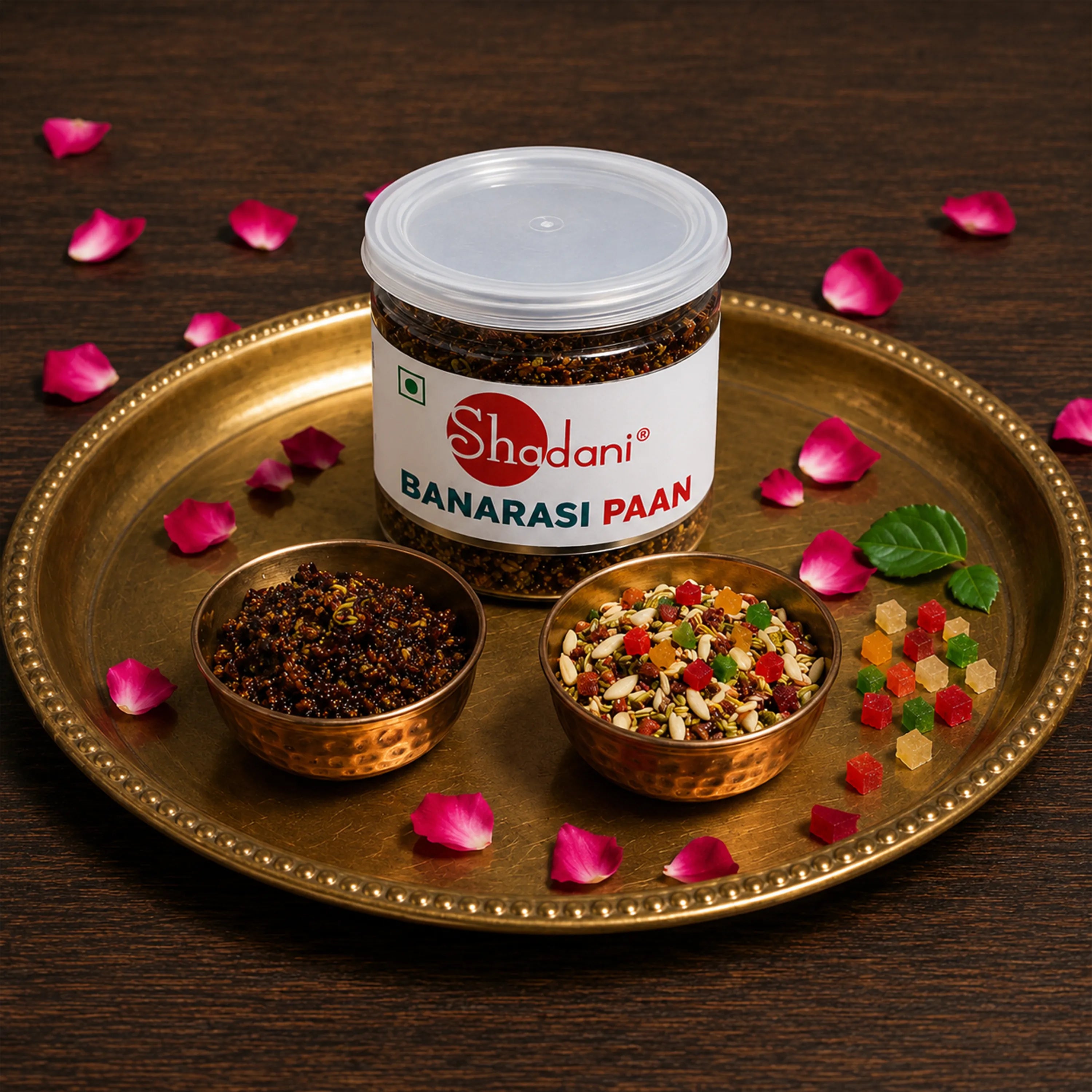 A jar of Shadani Banarasi Paan is displayed on a gold-colored tray with two small copper bowls containing the dried leaves and the prepared powder, surrounded by pink rose petals on a dark wooden background.