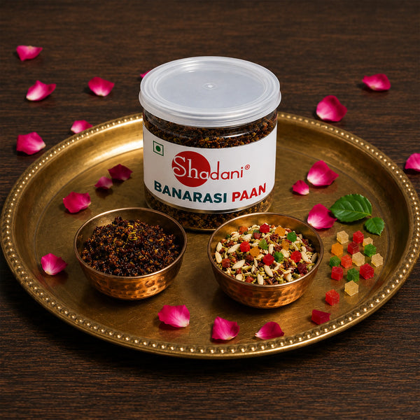 A jar of Shadani Banarasi Paan is displayed on a gold-colored tray with two small copper bowls containing the dried leaves and the prepared powder, surrounded by pink rose petals on a dark wooden background.