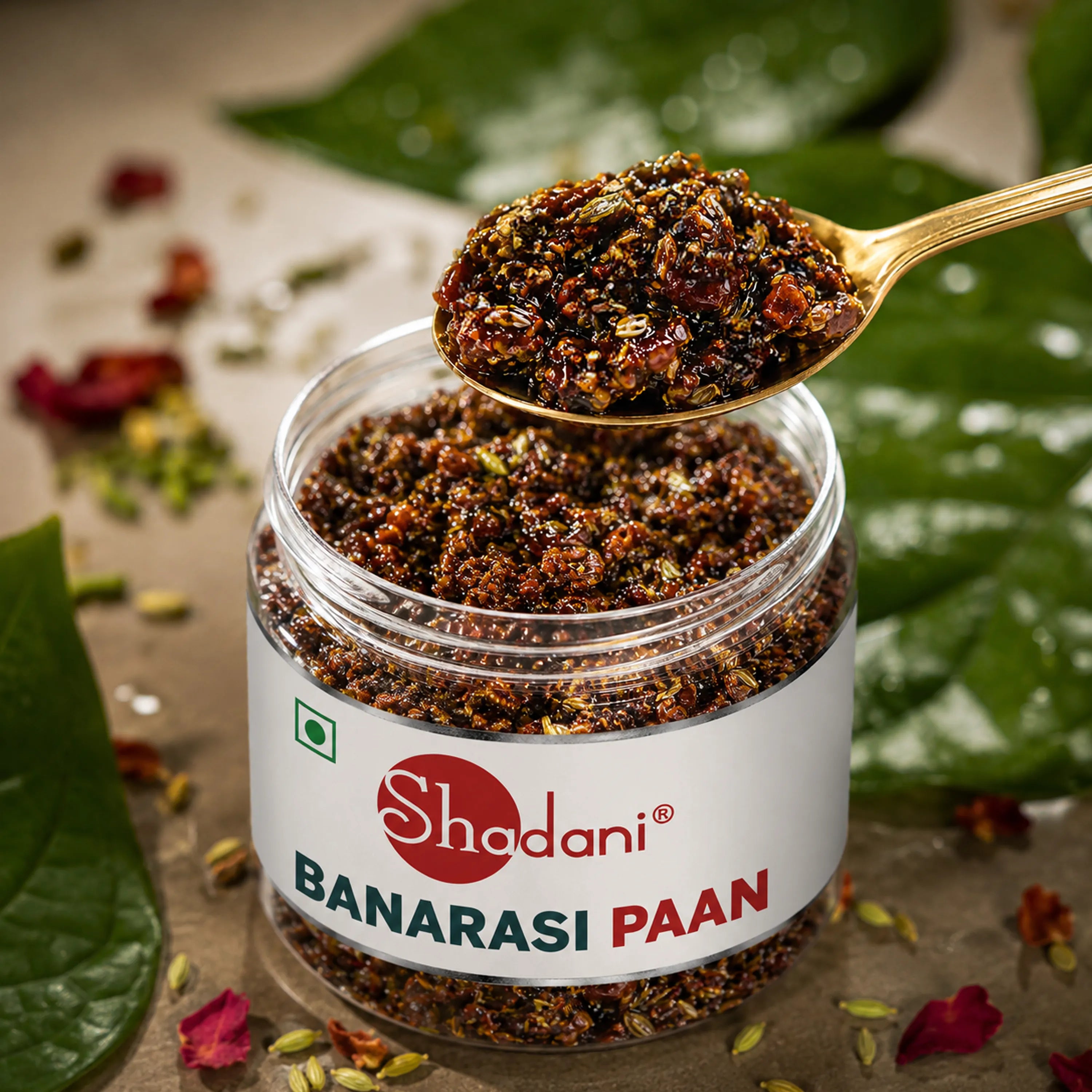 A clear plastic jar of Shadani Banarasi Paan with a white label, shown with a gold spoon holding a spoonful of the dark brown and red powder. The jar is surrounded by green leaves and scattered flower petals on a neutral surface.