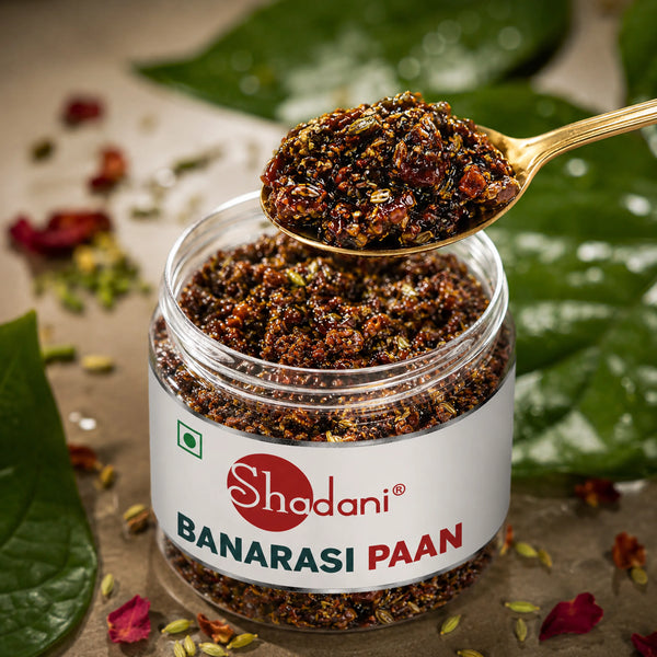 A clear plastic jar of Shadani Banarasi Paan with a white label, shown with a gold spoon holding a spoonful of the dark brown and red powder. The jar is surrounded by green leaves and scattered flower petals on a neutral surface.