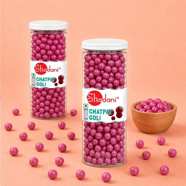 Two clear plastic jars of Shedani Chatpat Goli candy filled with pink goli balls, with a small wooden bowl of more goli candies, all arranged on a peach-colored background with scattered goli candies.
