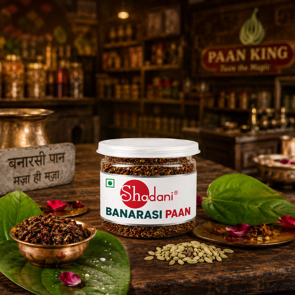 A jar of Shadani Banarasi Paan placed on a rustic wooden table with green leaves, small copper bowls, and loose capsules around it. The background is a warm, blurred traditional Indian setting with shelves and signage, including a visible Paan King sign.