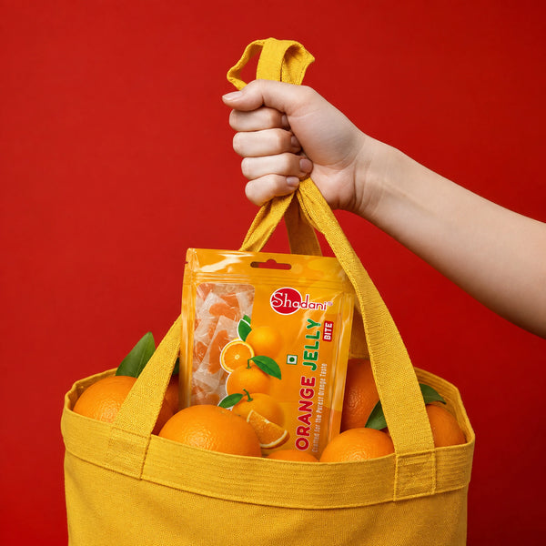 A hand holding the handle of a yellow canvas tote bag filled with oranges and a package of Shadani Orange Jelly gummies, set against a solid red background.