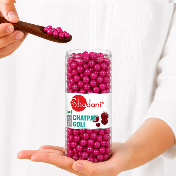 A person holds a clear cylindrical jar filled with bright magenta chatpatti goli beads, with a wooden spoon also holding some of the beads above the jar. The jar has a white label with the brand name 'Shedani' and the text 'CHATPATI GOLI'.