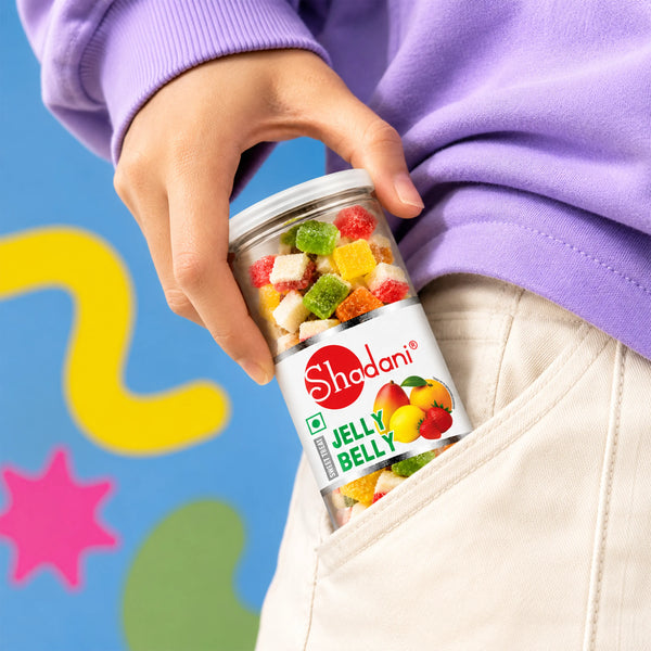 A person in a purple sweatshirt and cream pants holds a jar of Shadani Jelly Belly gummy candies in their pocket against a colorful blue background with abstract shapes.