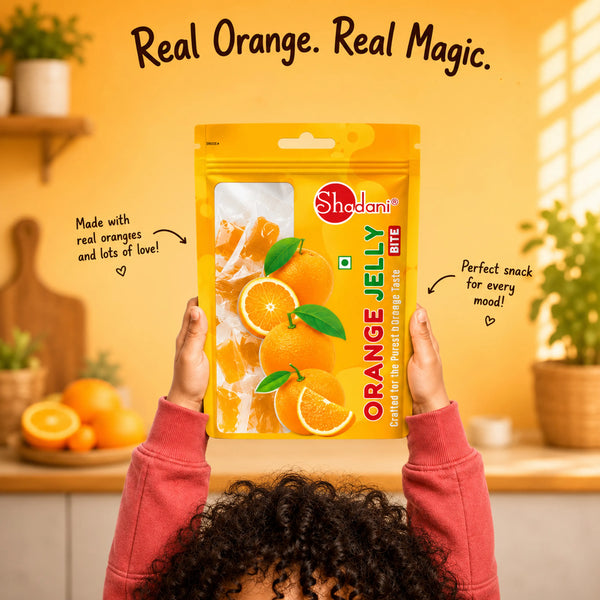 A person in a pink sweatshirt holds up a large package of Shedani Orange Jelly Gummies against a warm yellow kitchen background. The package features bright orange graphics and the Shadani brand logo.
