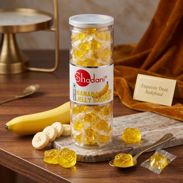 A clear plastic jar of Shadani Banana Jelly Balls sits on a wooden table with a marble slab, a banana, sliced bananas, gold spoons, and a small sign reading 'Exquisite Treat, Redeemed'. The jar is filled with translucent yellow jelly balls.