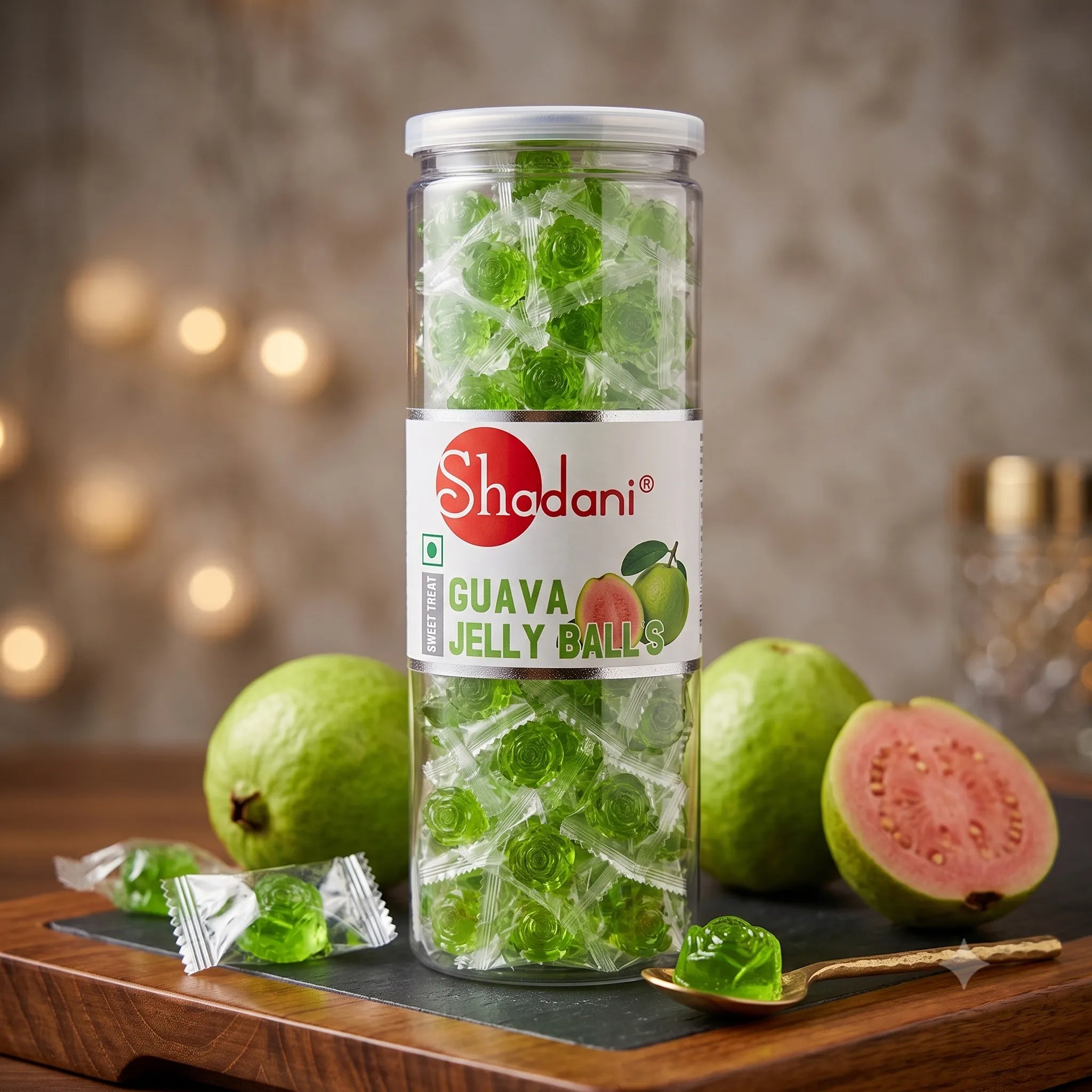 A clear cylindrical jar of Shadani Guava Jelly Balls filled with green translucent jelly balls, standing on a dark tray with whole and sliced guava fruits, loose wrapped pieces, and a spoon around it on a wooden surface.