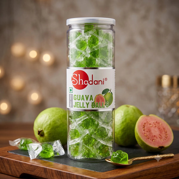 A clear cylindrical jar of Shadani Guava Jelly Balls filled with green translucent jelly balls, standing on a dark tray with whole and sliced guava fruits, loose wrapped pieces, and a spoon around it on a wooden surface.