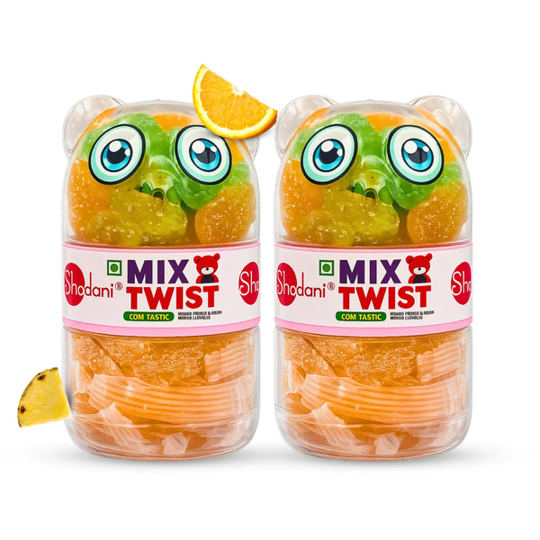 Two clear plastic jars of Shadani Twist Comb Tasty gummy candy, shaped like cartoon frog faces with green and orange gummies inside, shown on a white background with orange and yellow garnish slices.